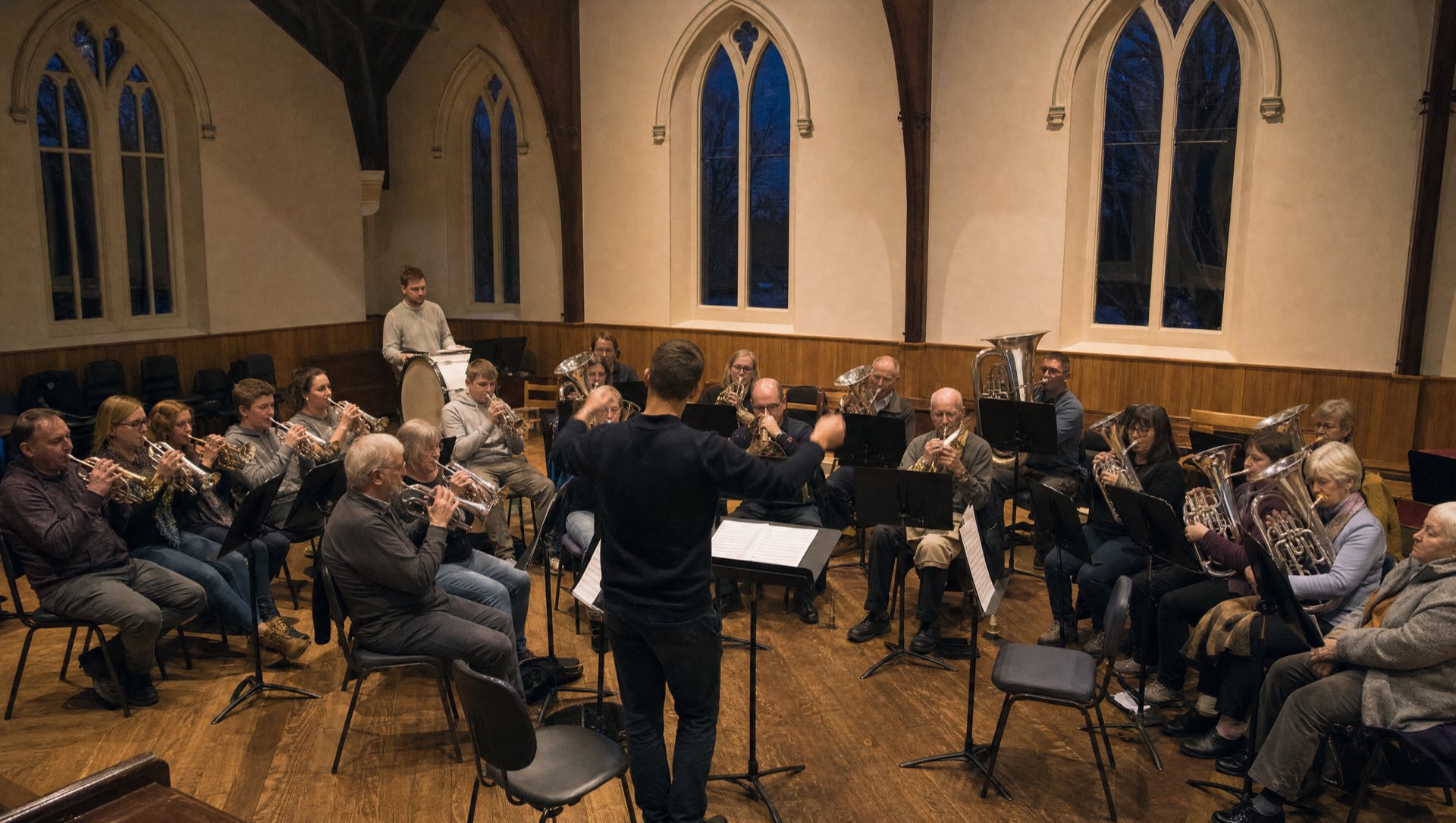 Garforth Brass in rehearsal: a semicircle of brass players and music stands in a church hall, the conductor's arms raised.
